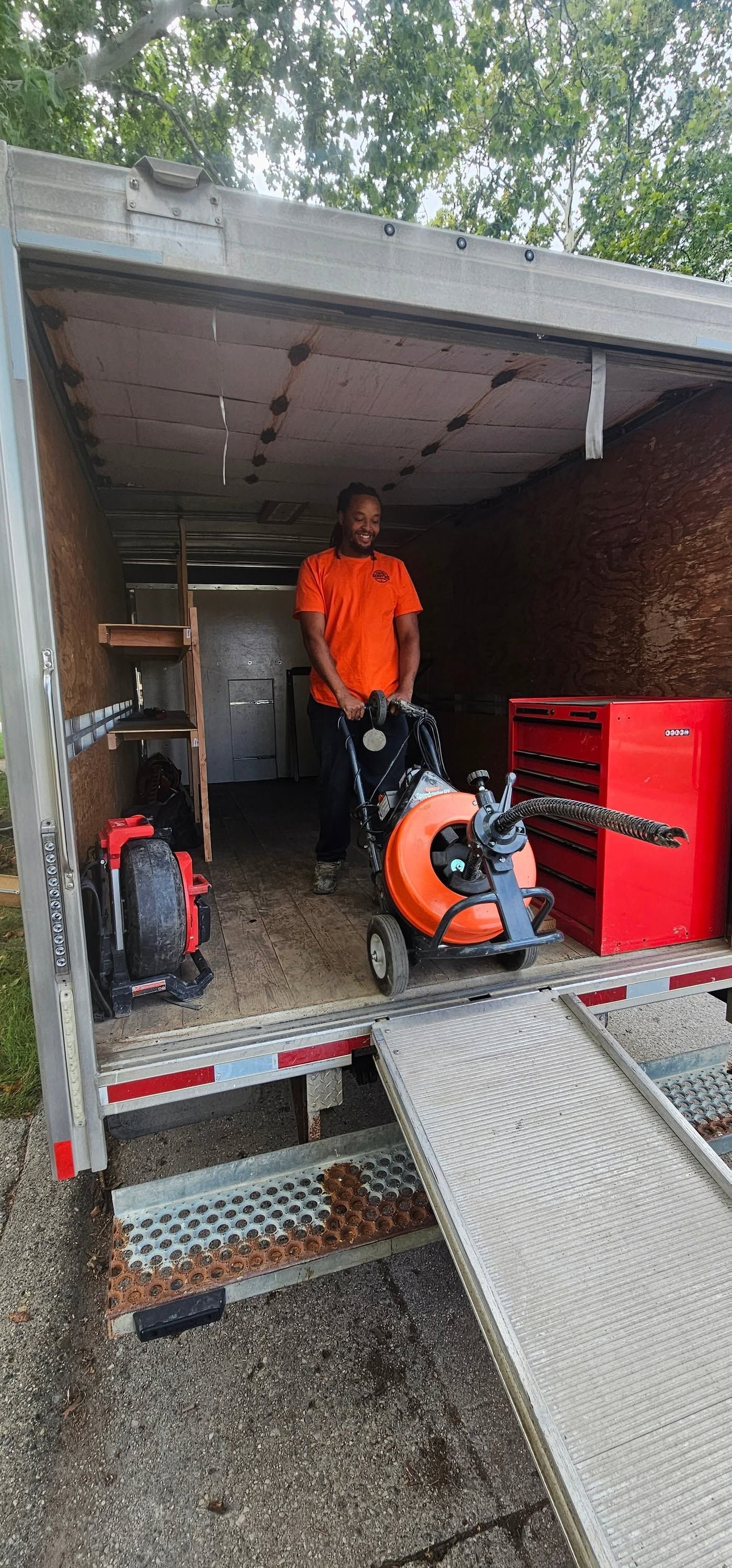 Person in orange shirt standing in a garage with a leaf blower, lawn tools, and a red tool chest.