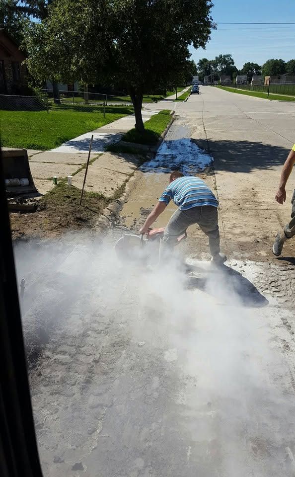 Worker jackhammering sidewalk, sending up dust beside a suburban street.