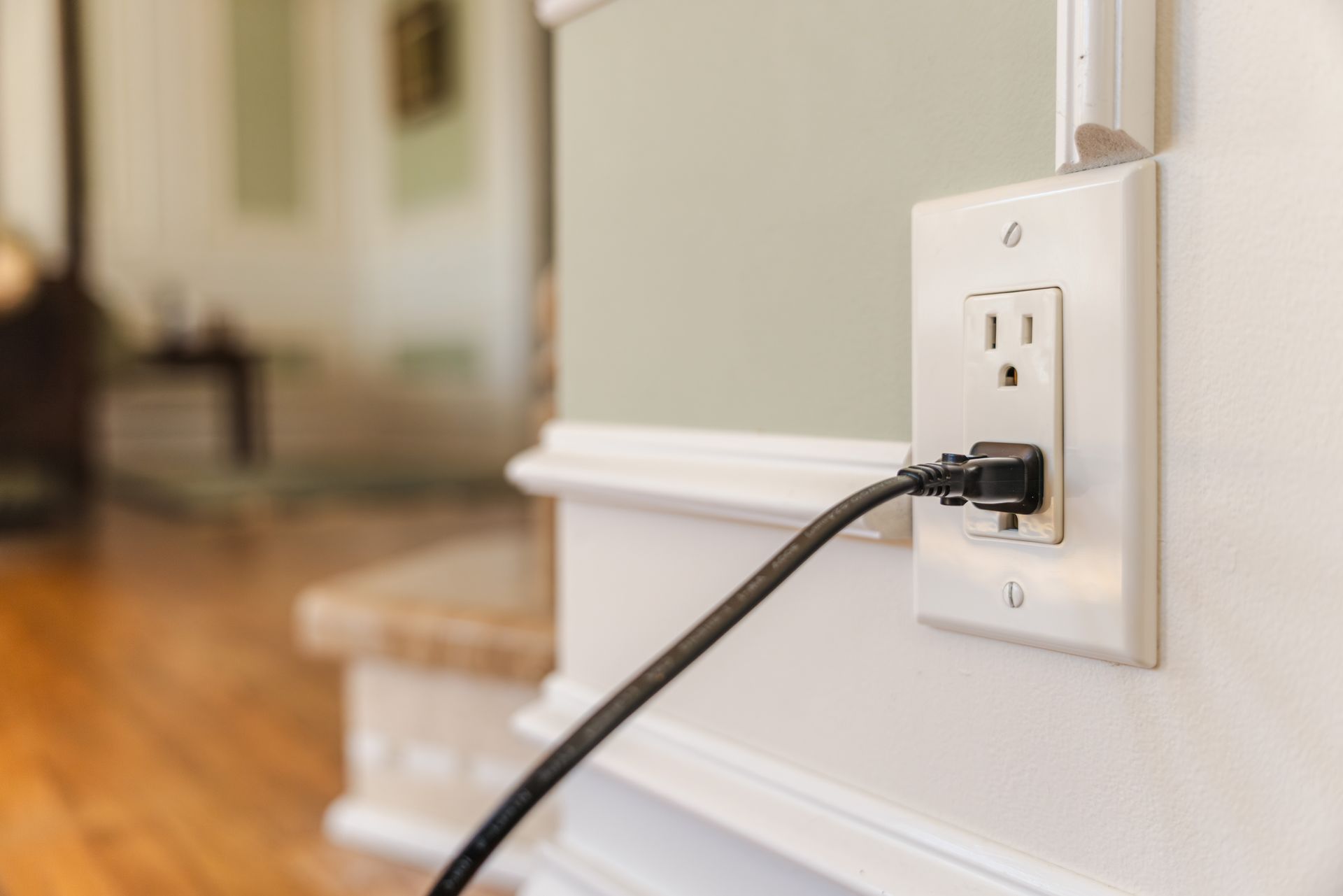 Black charger plugged into a white wall outlet near a staircase in a home interior