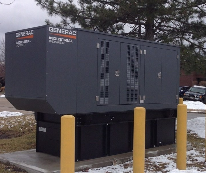 Large dark gray Generac industrial generator outside, surrounded by yellow bollards and snow.