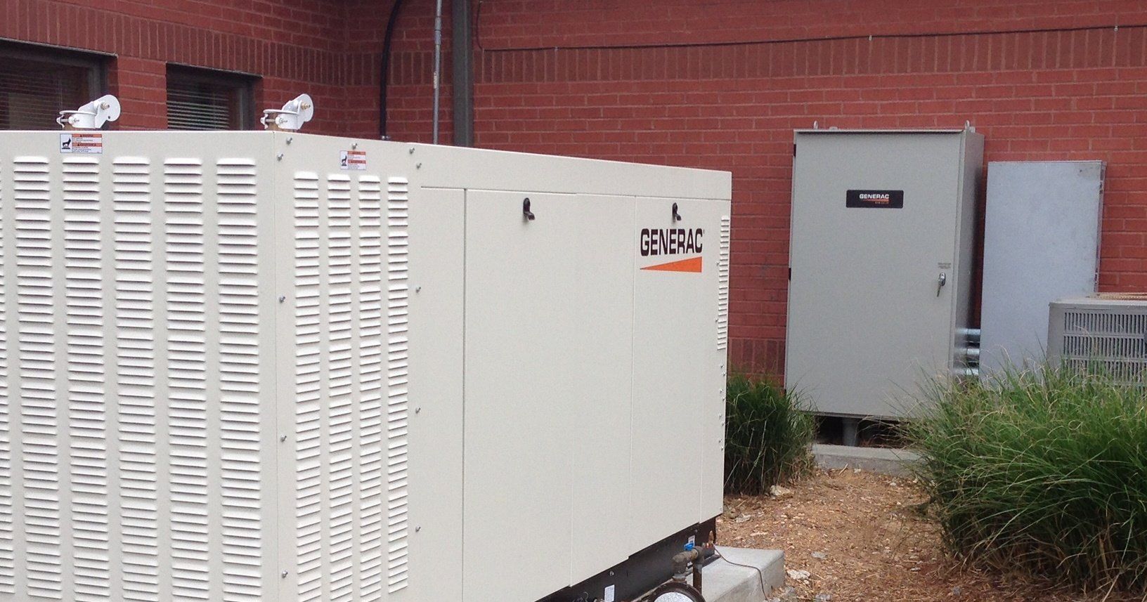 Two white utility cabinets beside a brick building, with a gray electrical box and tall grass nearby