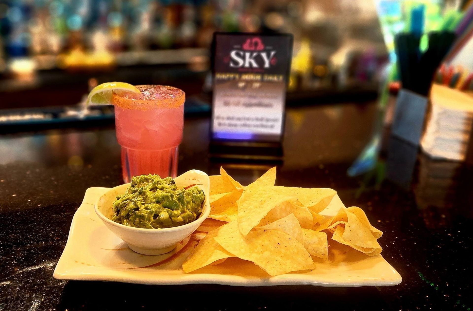 Cocktail, guacamole, and tortilla chips on a tray at a bar with a blurred SKY sign in back