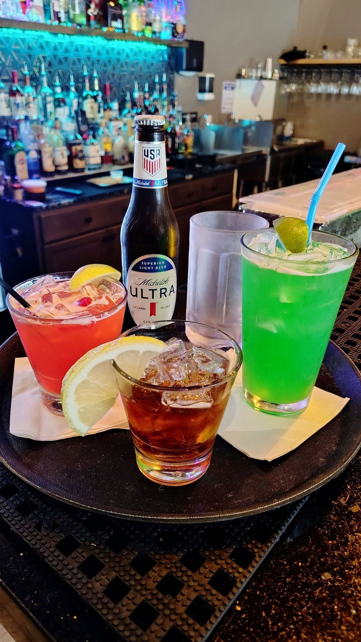 Two colorful cocktails and a whiskey shot on a bar table, with a liquor bottle and bar shelves behind.