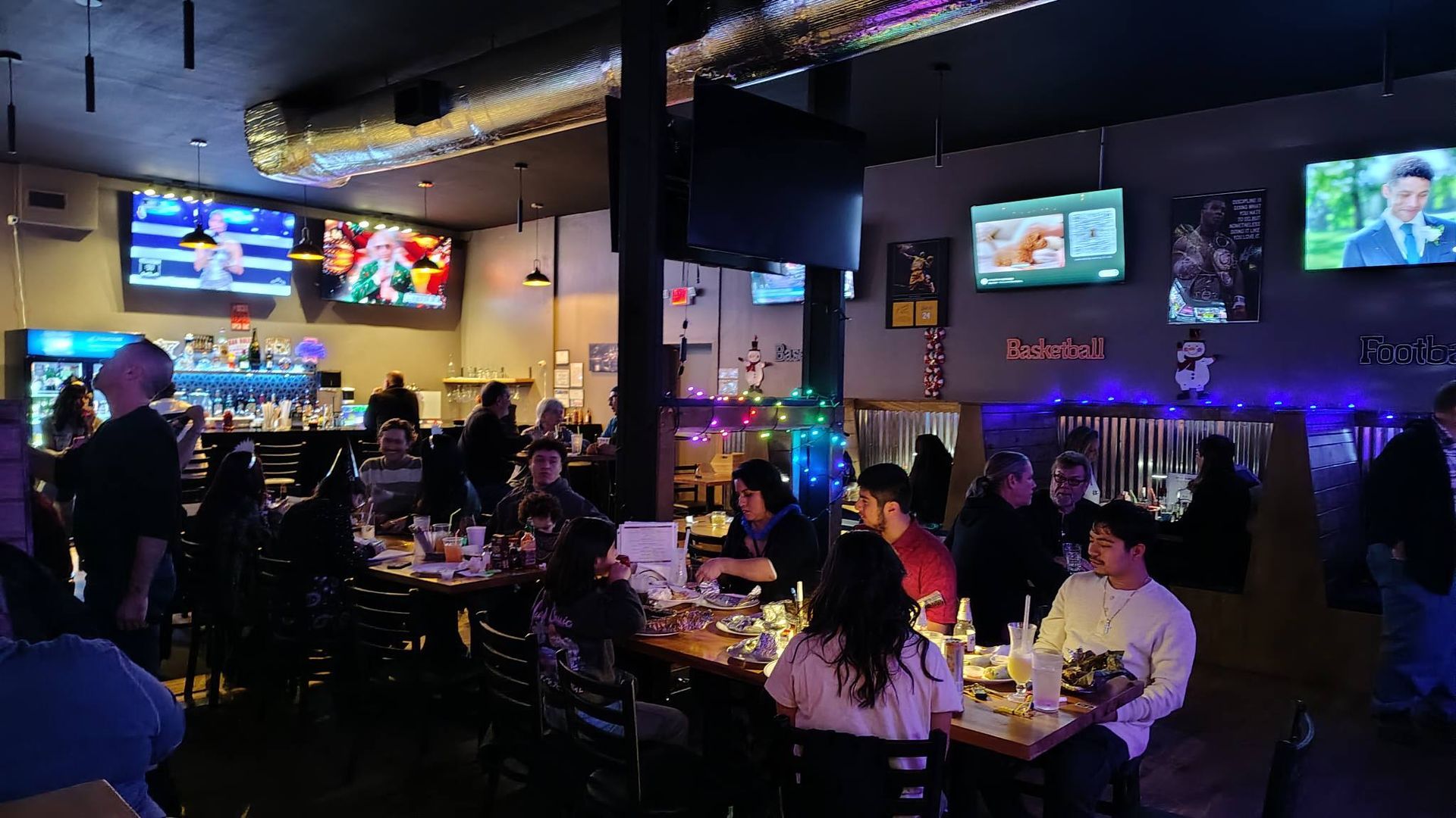 Dimly lit sports bar with people dining at tables and TVs showing games in the background.
