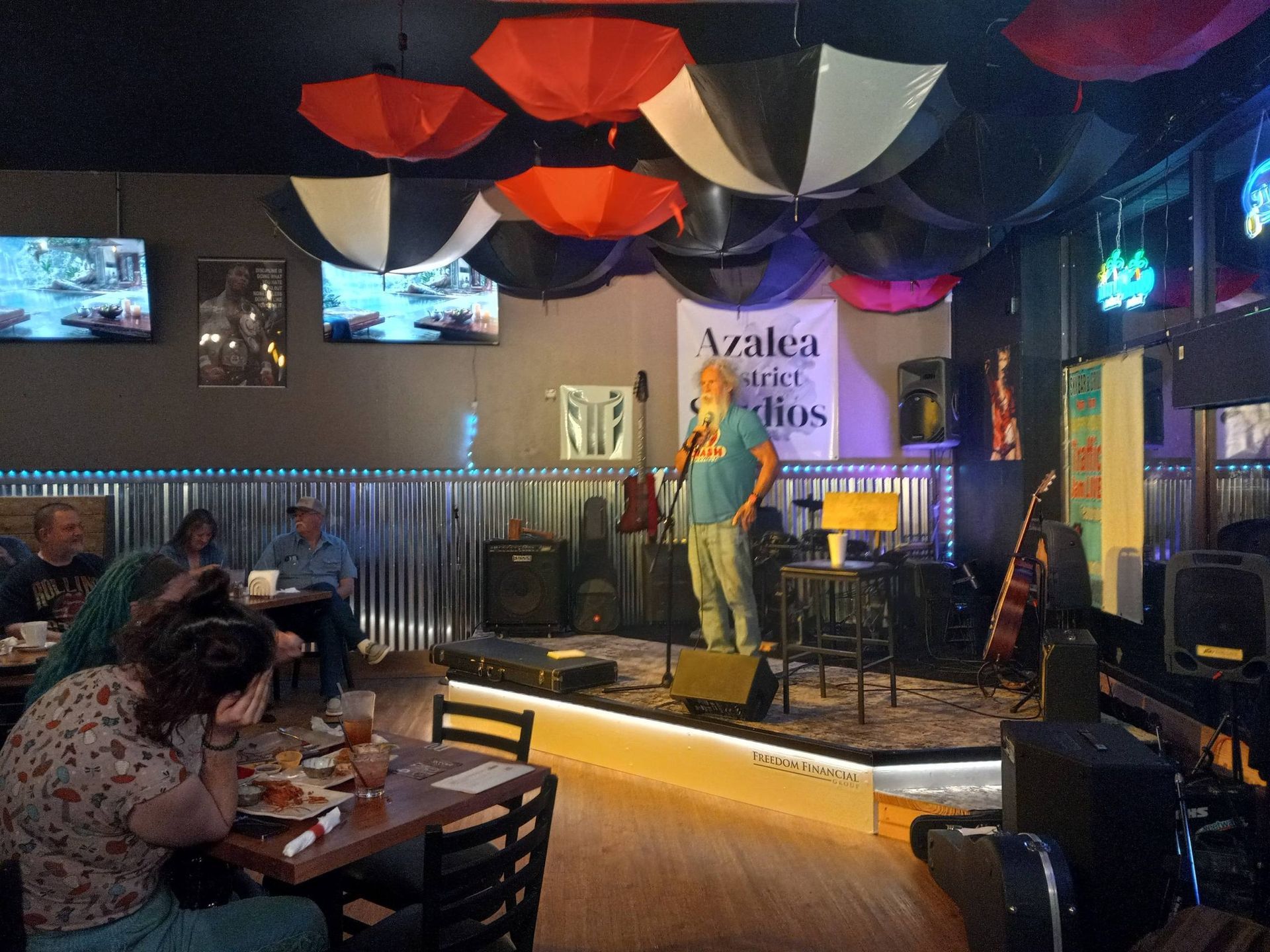 Small stage in a café with a singer performing under colorful ceiling fabric while patrons sit at tables.