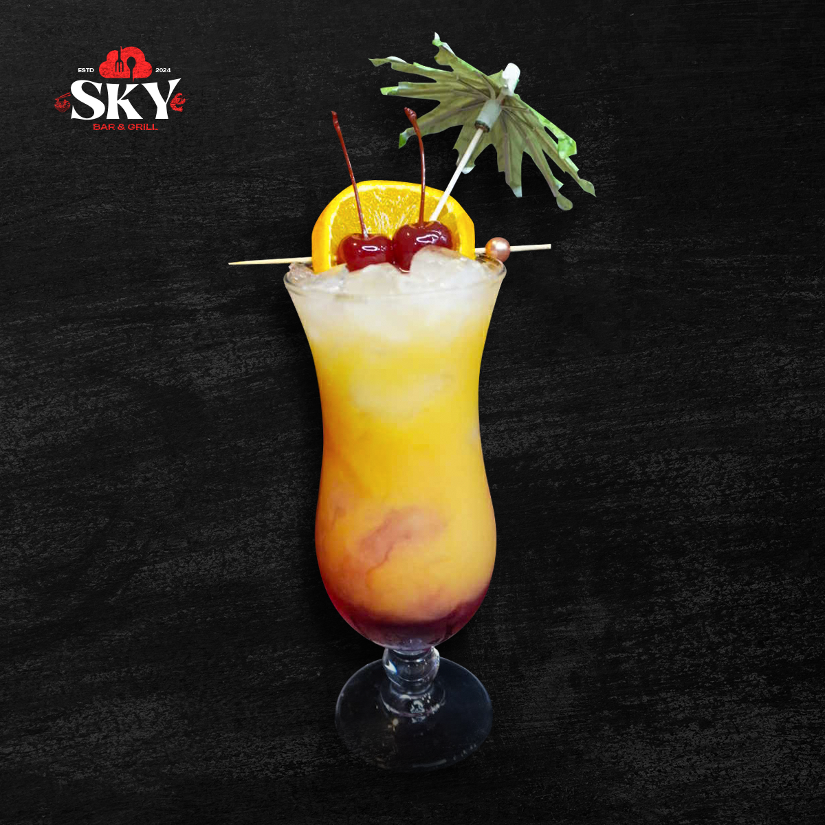 Yellow-orange layered cocktail with cherries, pineapple, and mint garnish in a stemmed glass on a black background