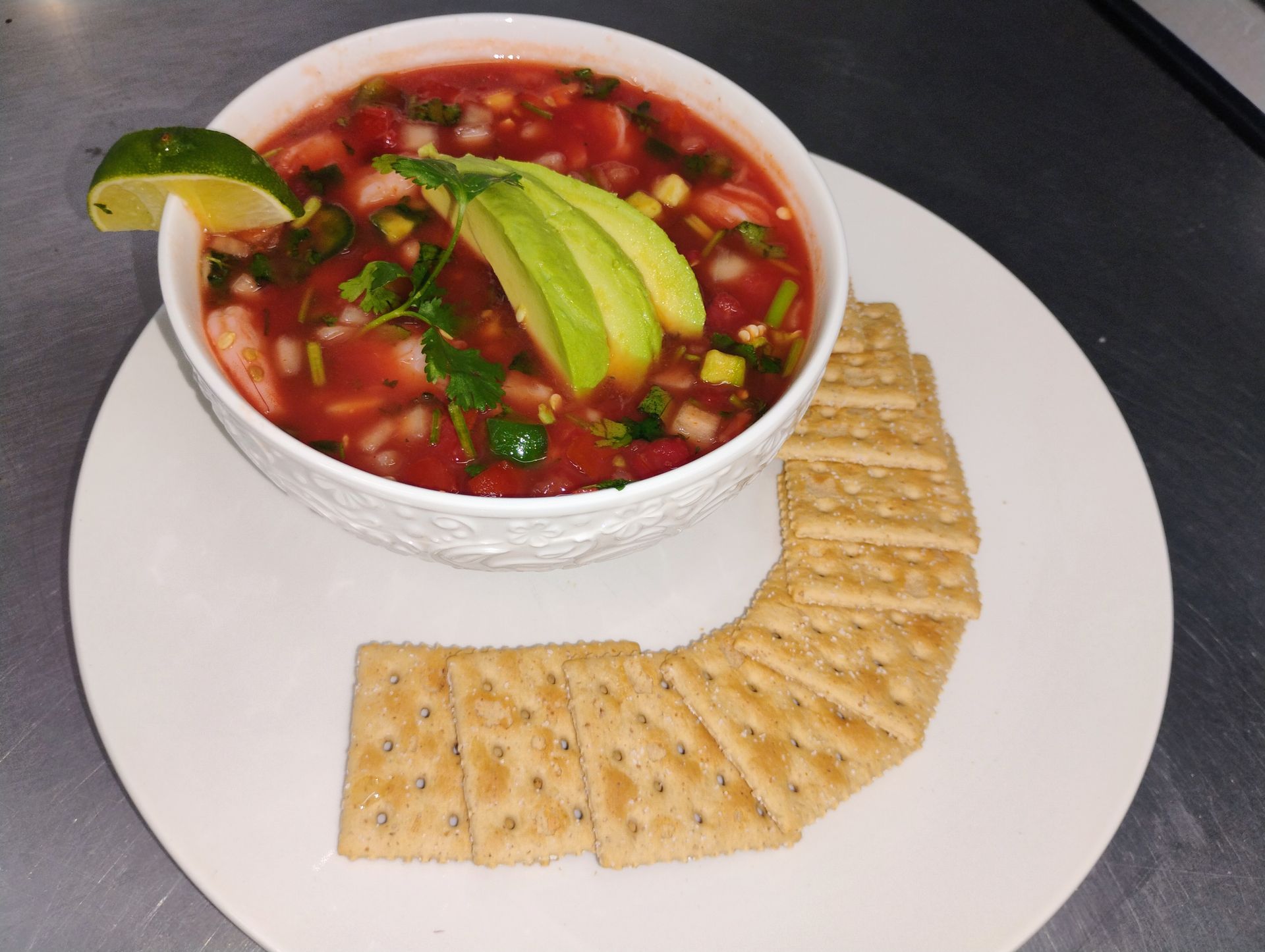 Bowl of red salsa topped with avocado, served with crackers on a white plate