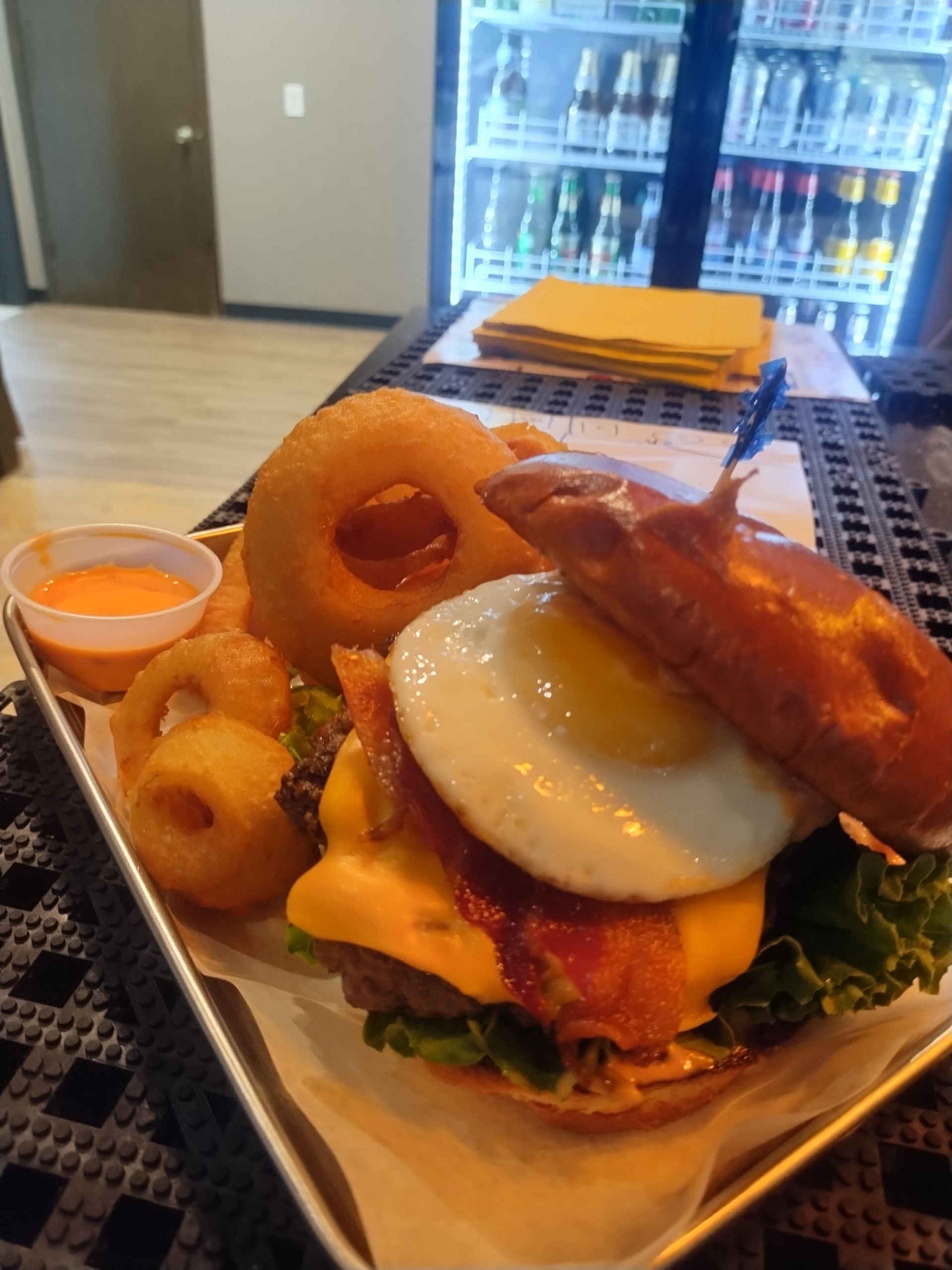 Bacon burger with fried egg, onion rings, and dipping sauce on a tray in a casual restaurant