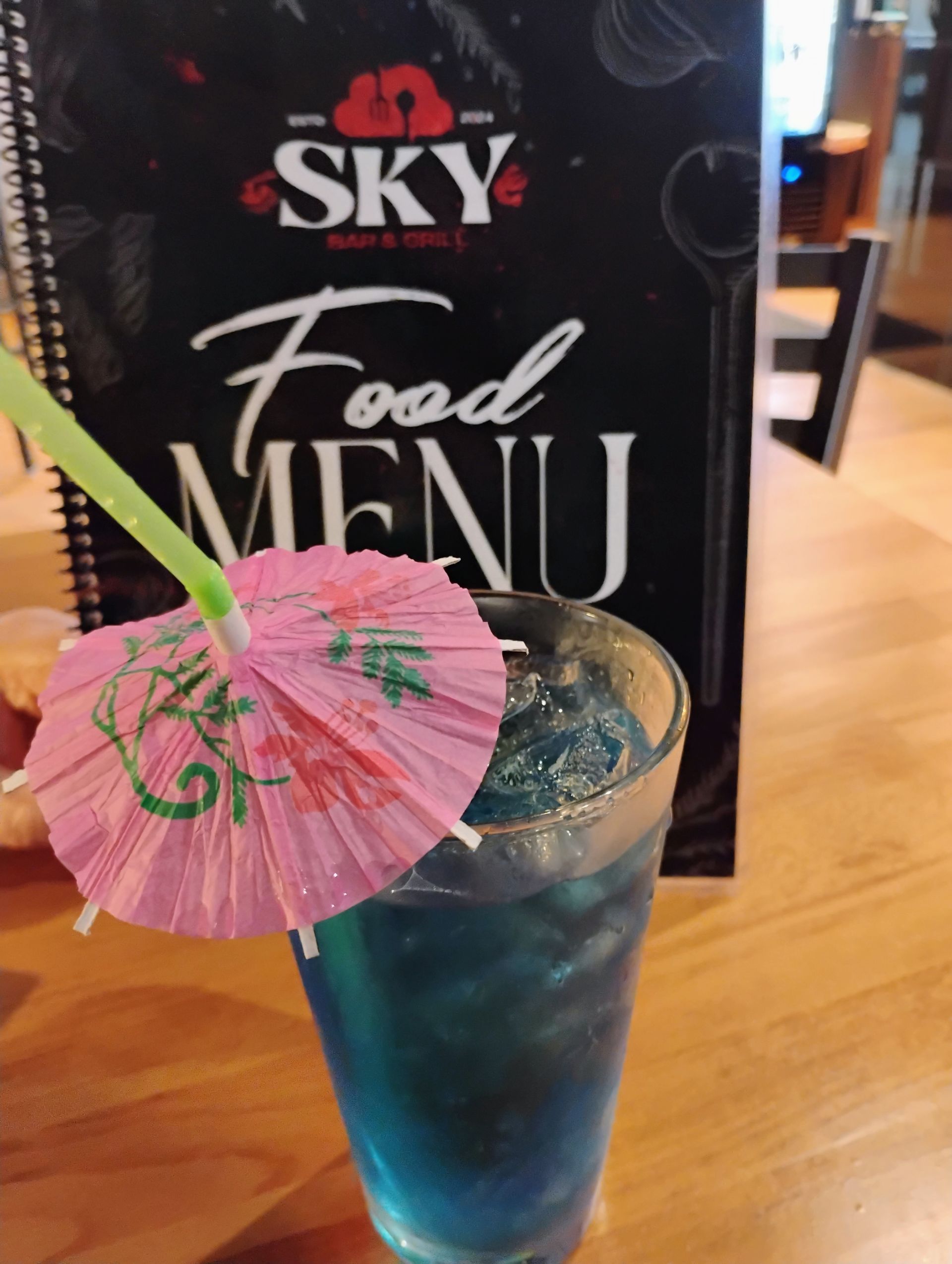 Blue cocktail with pink paper umbrella on a wooden table in front of a food menu sign.
