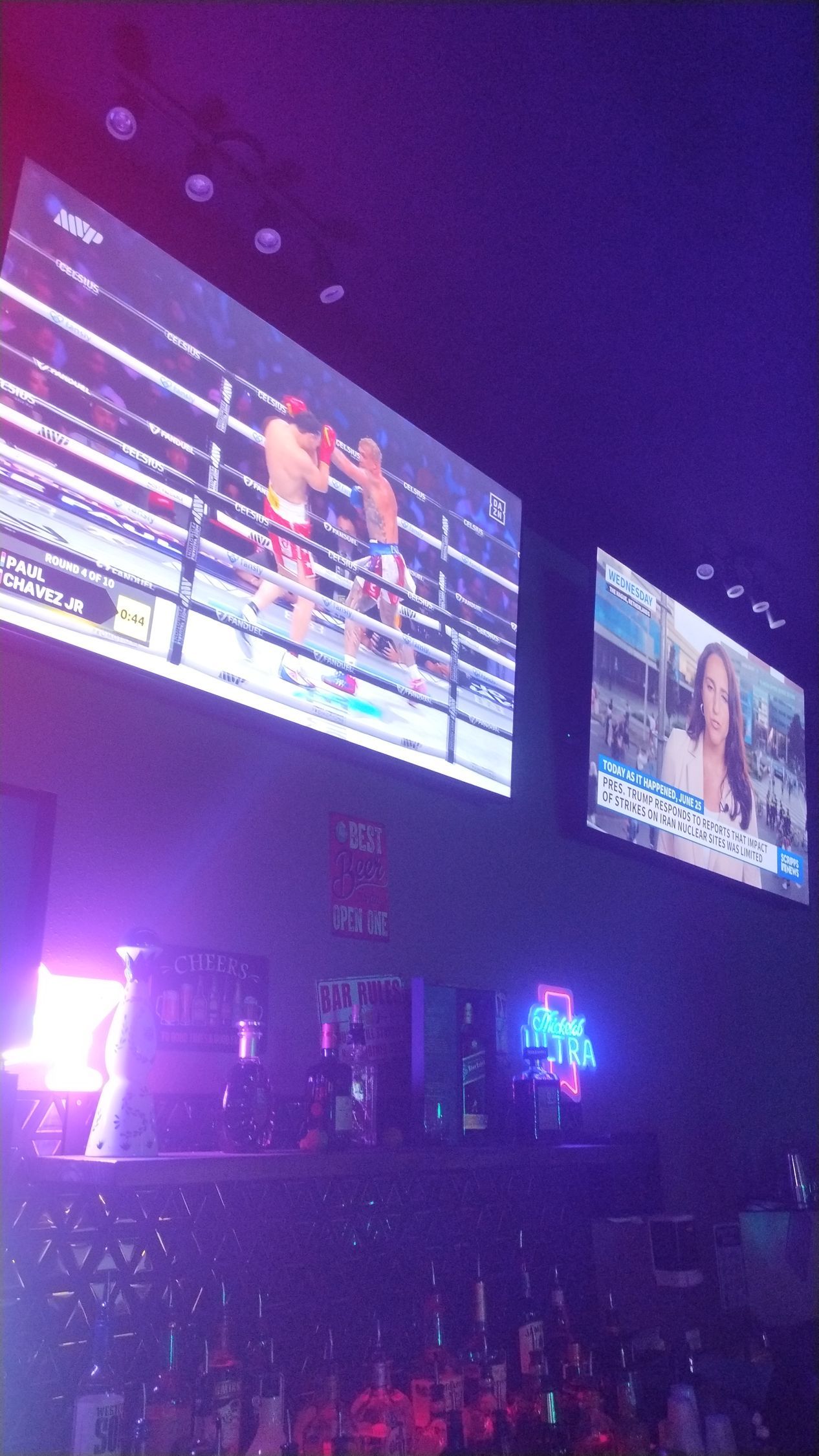 Two wall-mounted TVs in a dim, purple-lit room above shelves of bottles.