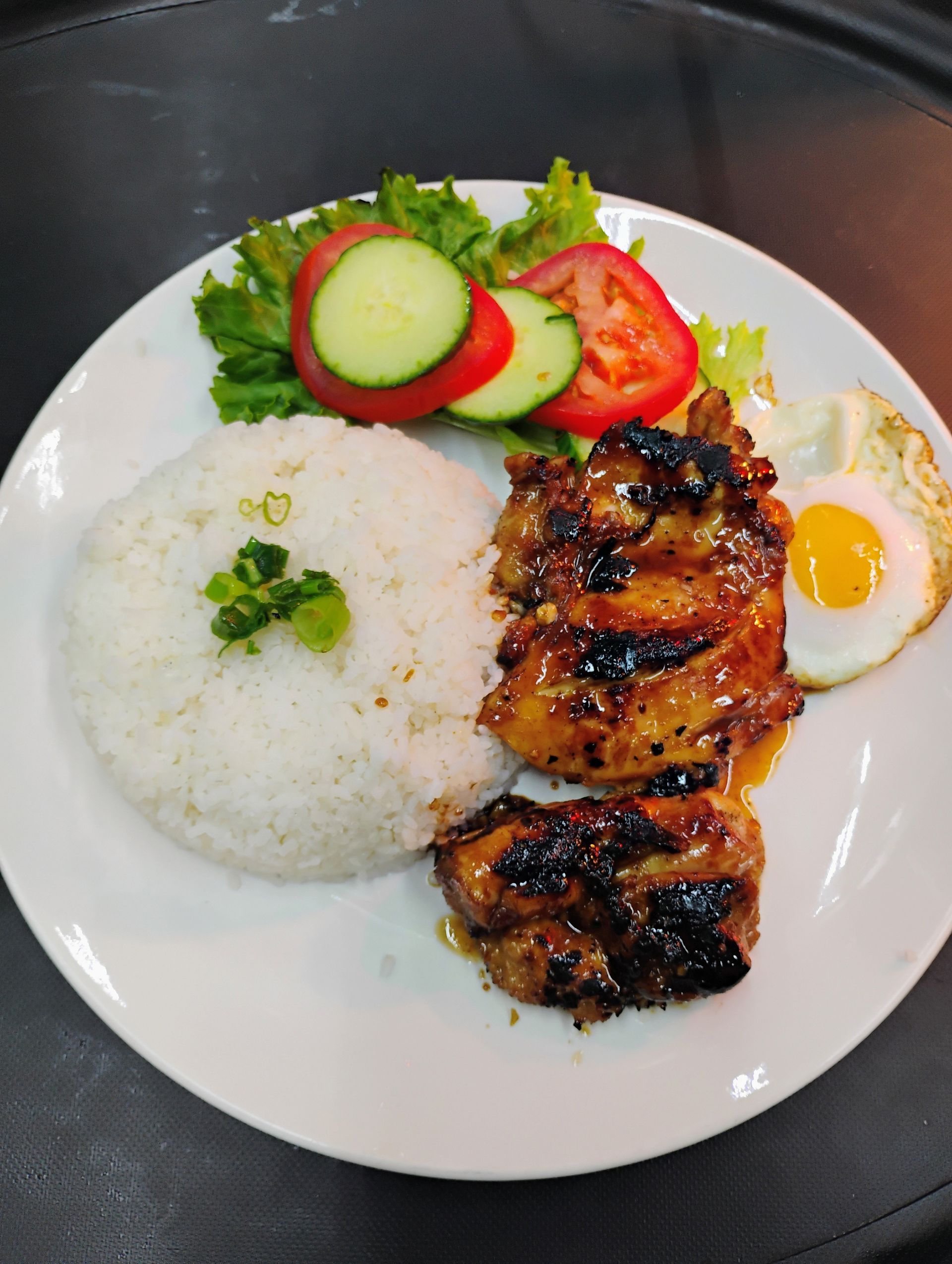 Grilled chicken, rice, fried egg, and sliced cucumber-tomato salad on a white plate