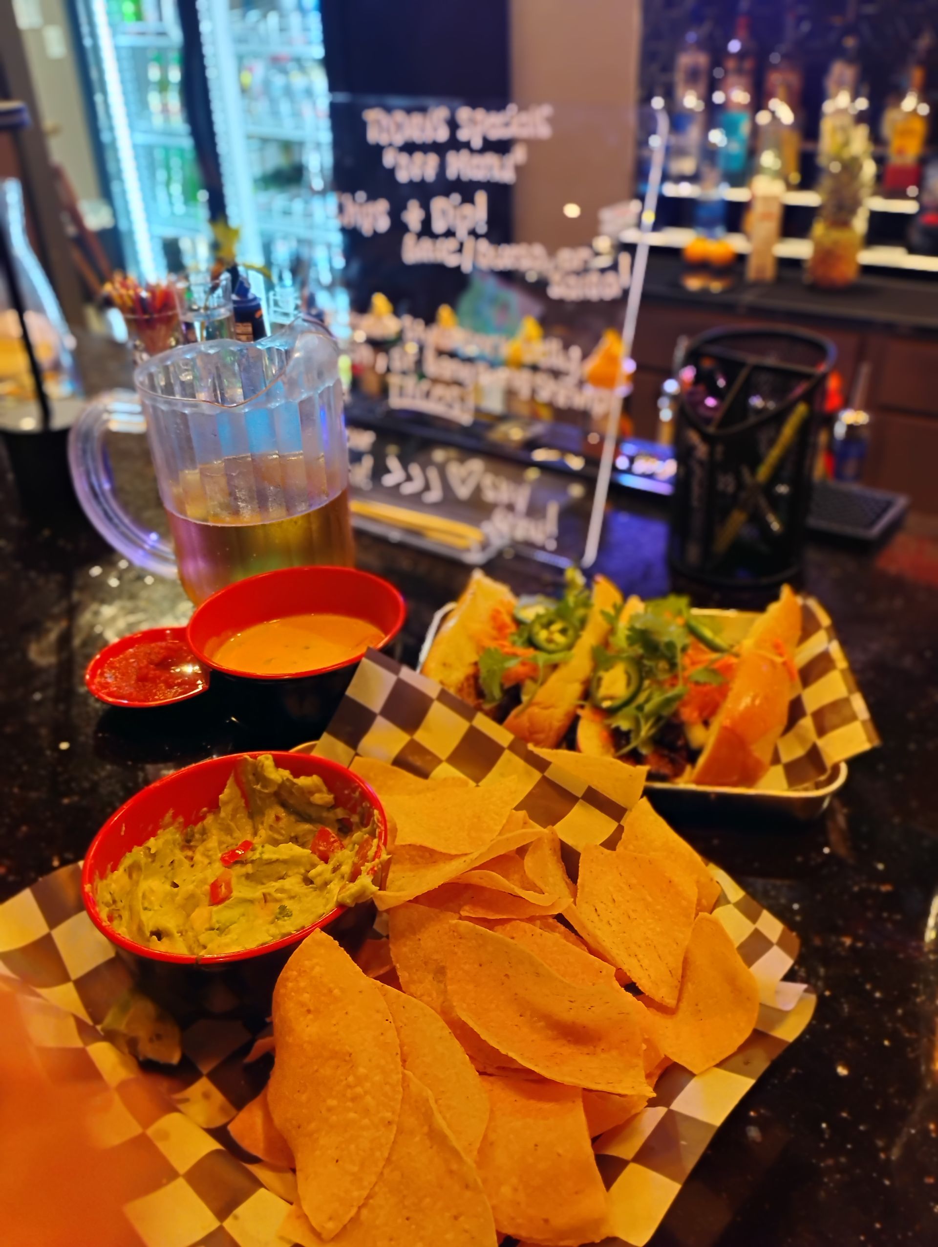 Three bowls of salsa, guacamole, and tortilla chips on a checkered tray at a bar counter