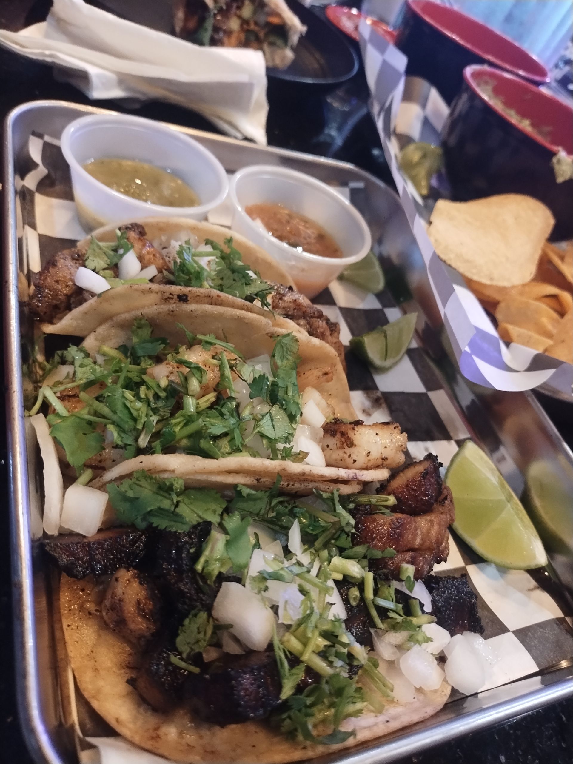 Taco platter with cilantro, onions, lime wedges, salsa cups, and tortilla chips on a checkered tablecloth