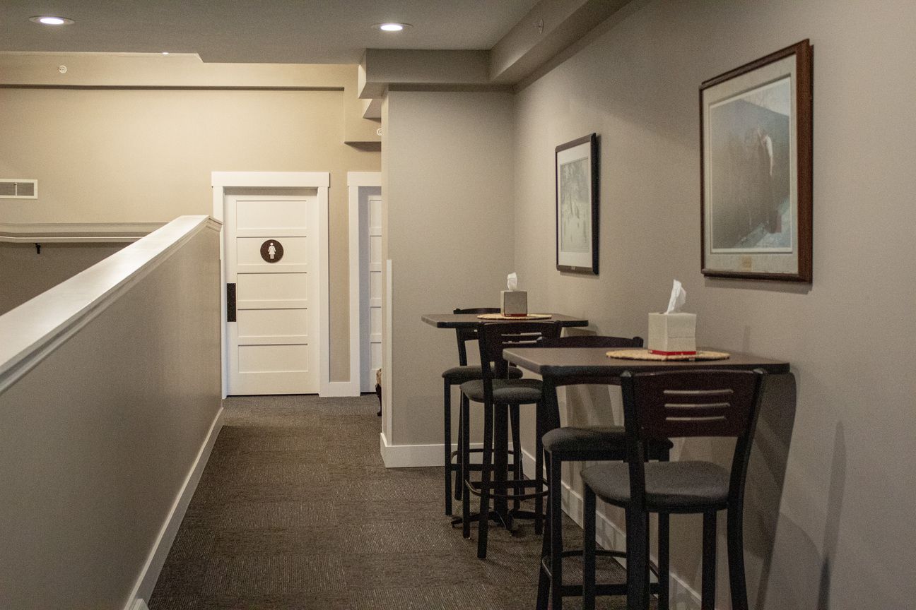 Empty hallway with small tables and chairs beside framed wall art, in a beige office or hotel lobby.