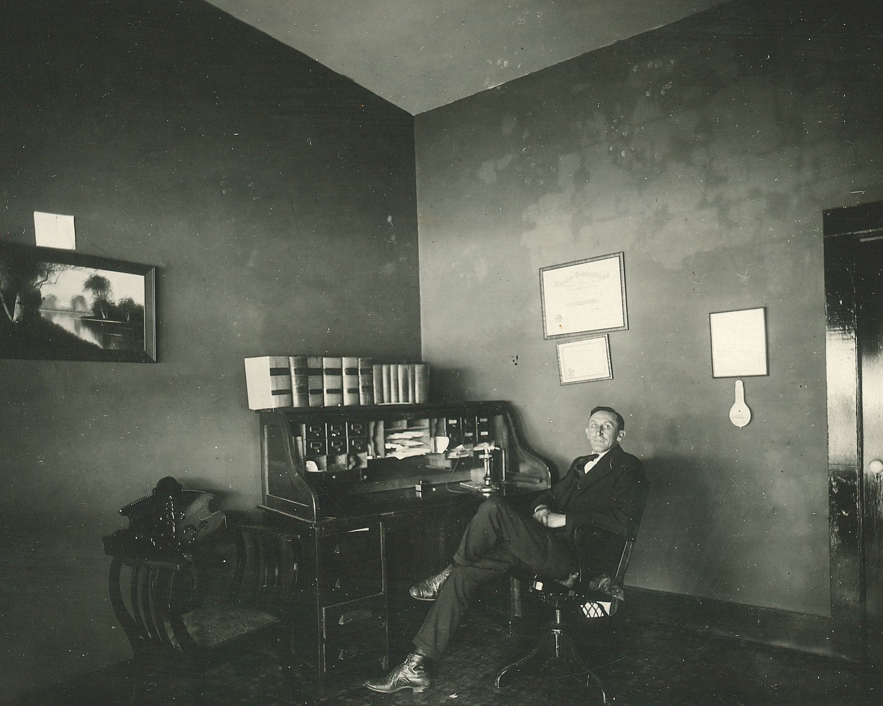 A man sits in a dim room with bookshelves, a framed picture, and wall lights.