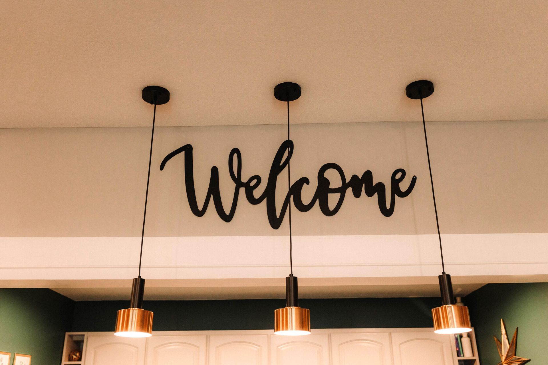 Black cursive “Welcome” sign above three hanging pendant lights indoors