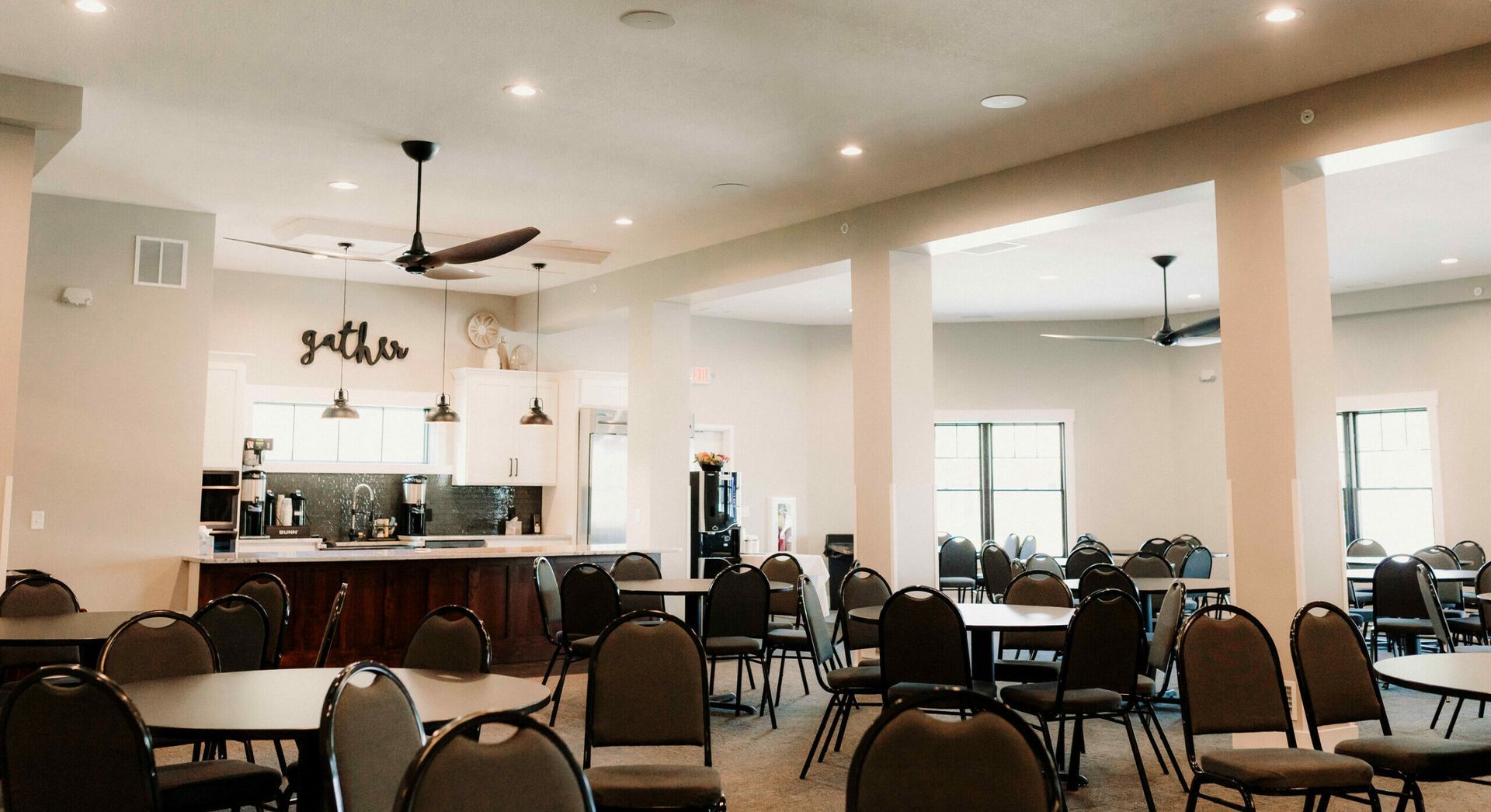 Empty banquet hall with round tables and chairs, ceiling fans, and a white reception counter in back