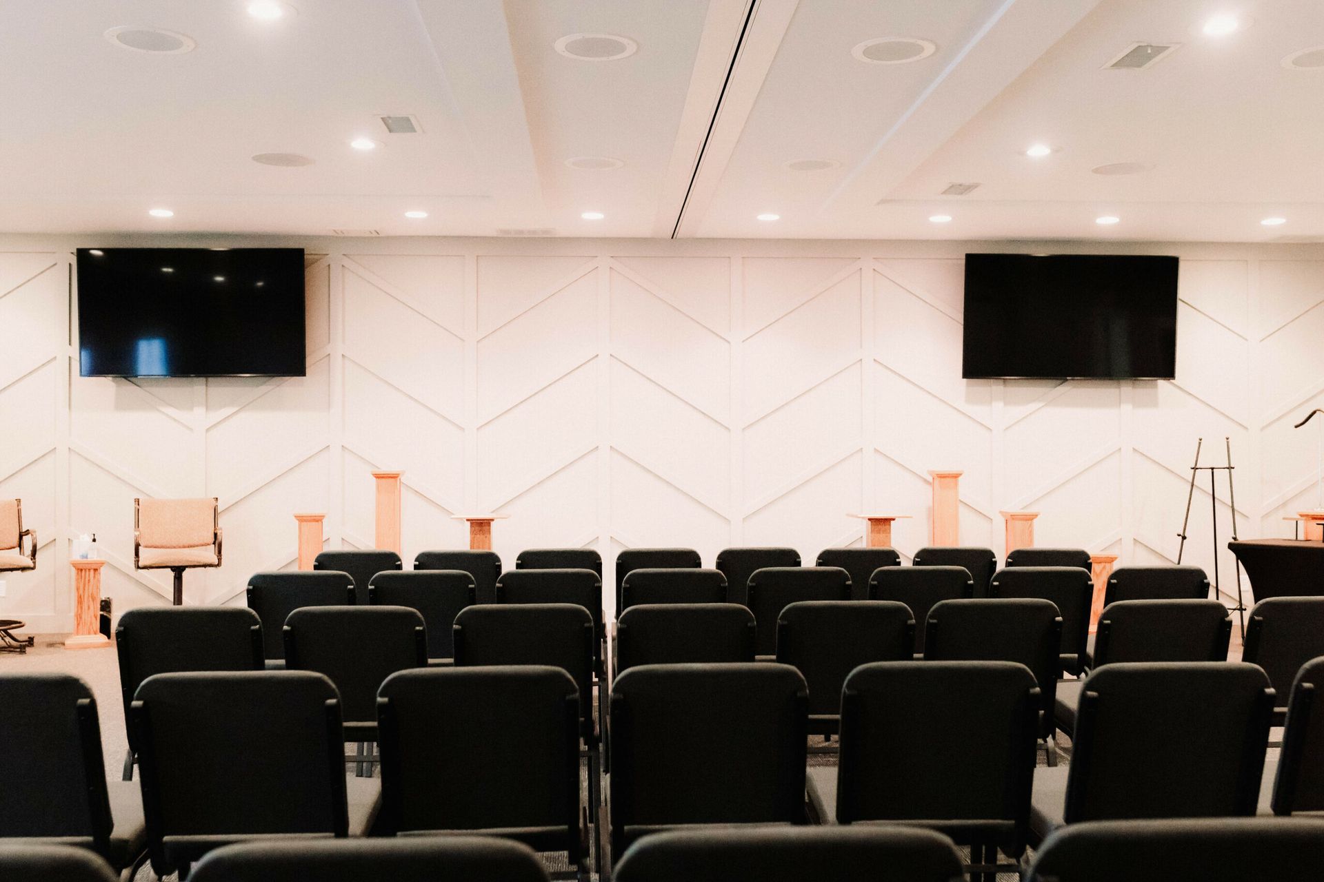 Empty conference room with rows of black chairs, podiums, and two wall-mounted screens at the front