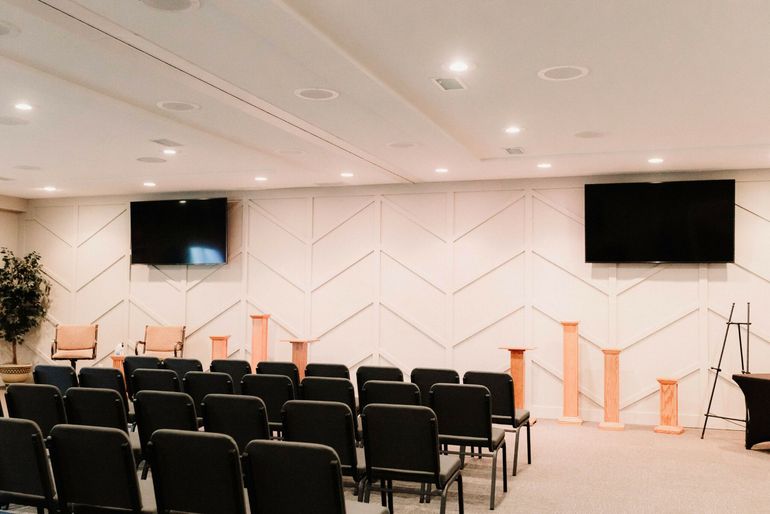 Empty conference room with rows of black chairs, podiums, and two wall-mounted screens on a white geometric wall