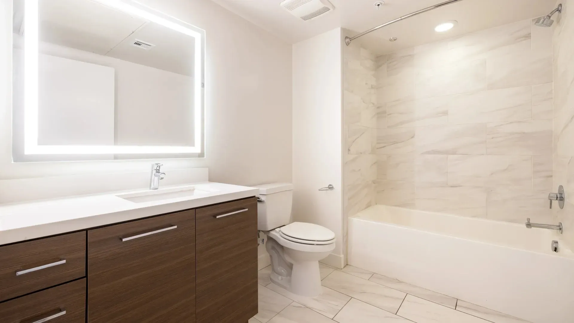 Modern bathroom with a large illuminated mirror, vanity, toilet, and bathtub with marble tile shower surround at Next on Sixth Los Angeles