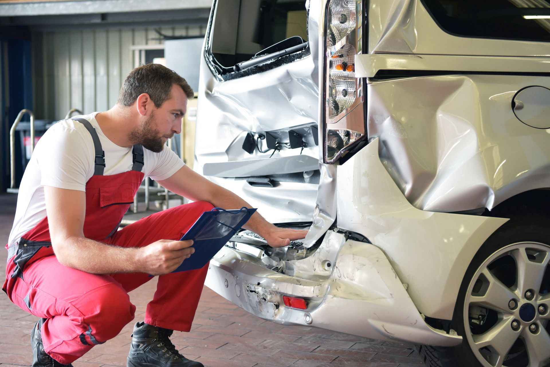 A technician in red work clothes holds a clipboard while inspecting the damaged rear of a white car in a garage.