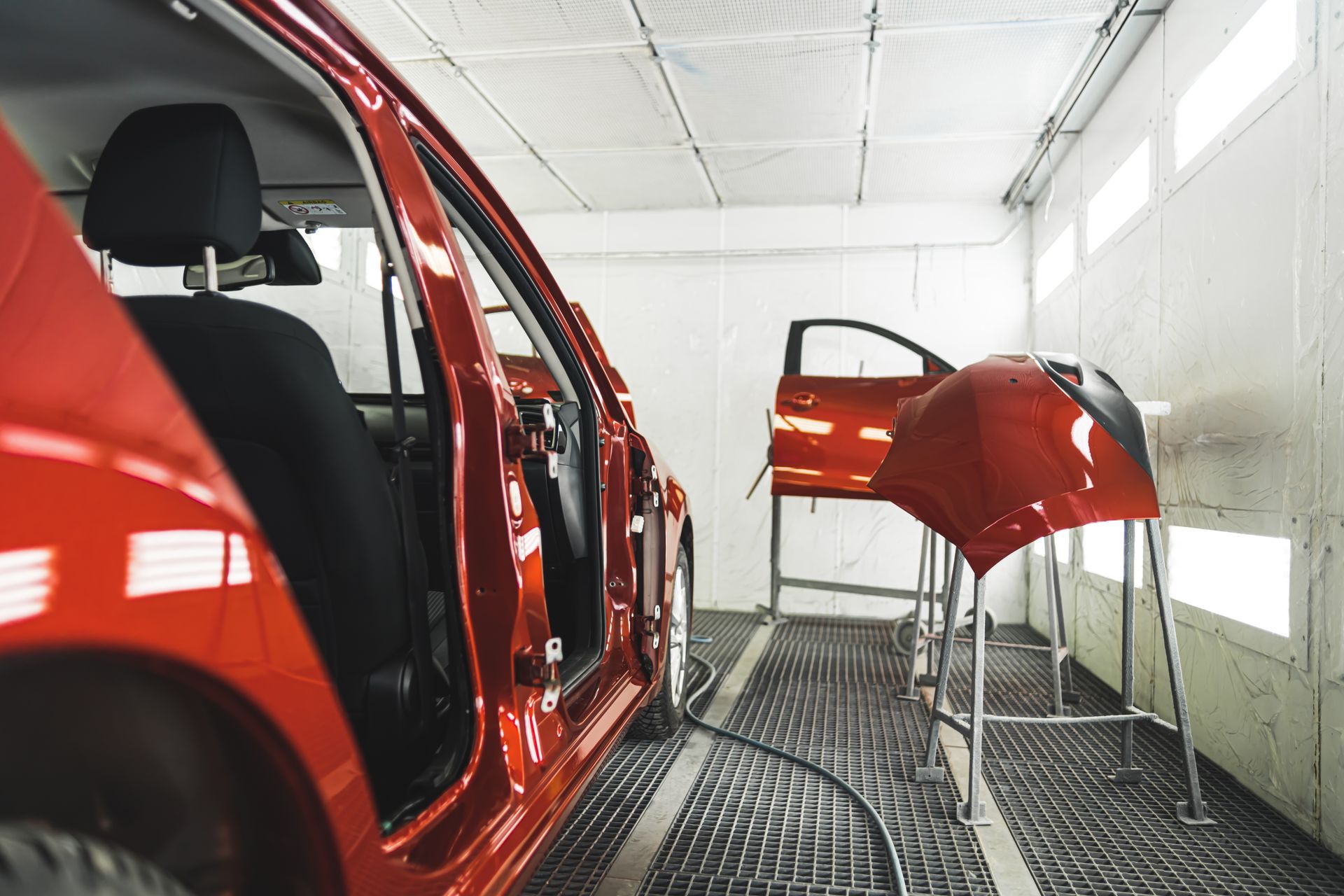 A red car body and detached parts in a brightly lit industrial paint booth.