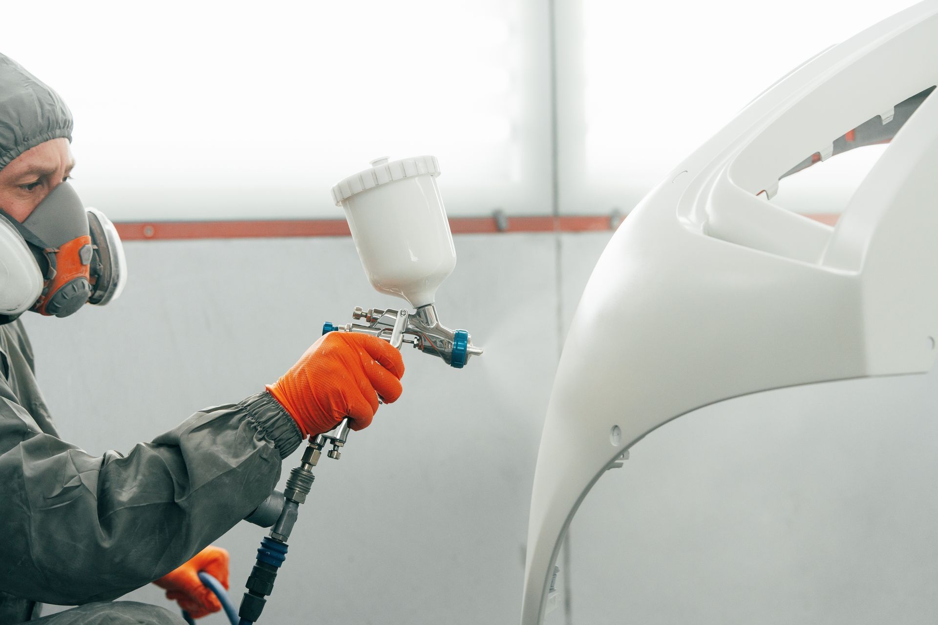 A person in protective gear and a respirator sprays white paint onto a car bumper with a spray gun.