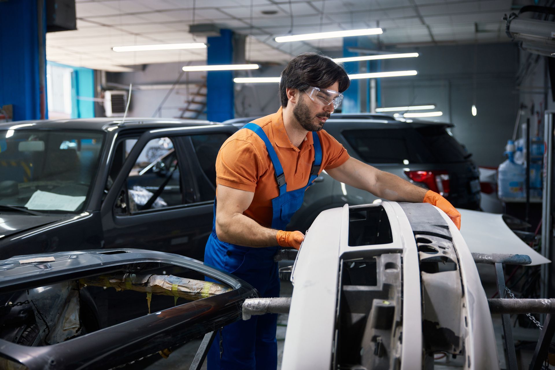 A technician in an orange shirt and blue overalls works on a car bumper in an auto repair shop.