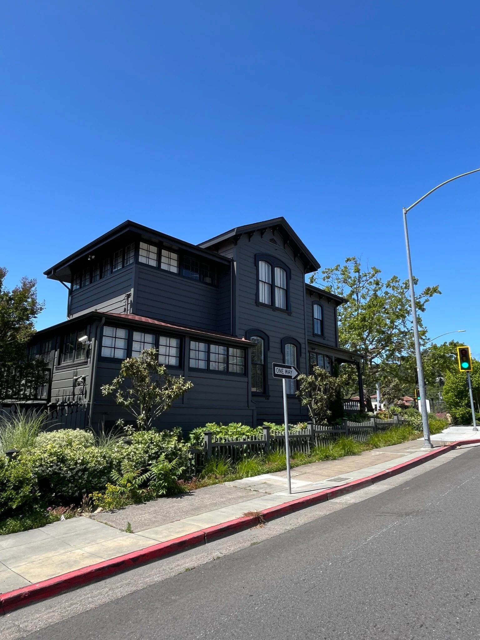 Dark gray two-story house with white-framed windows, surrounded by greenery and a street, under a bright blue sky.