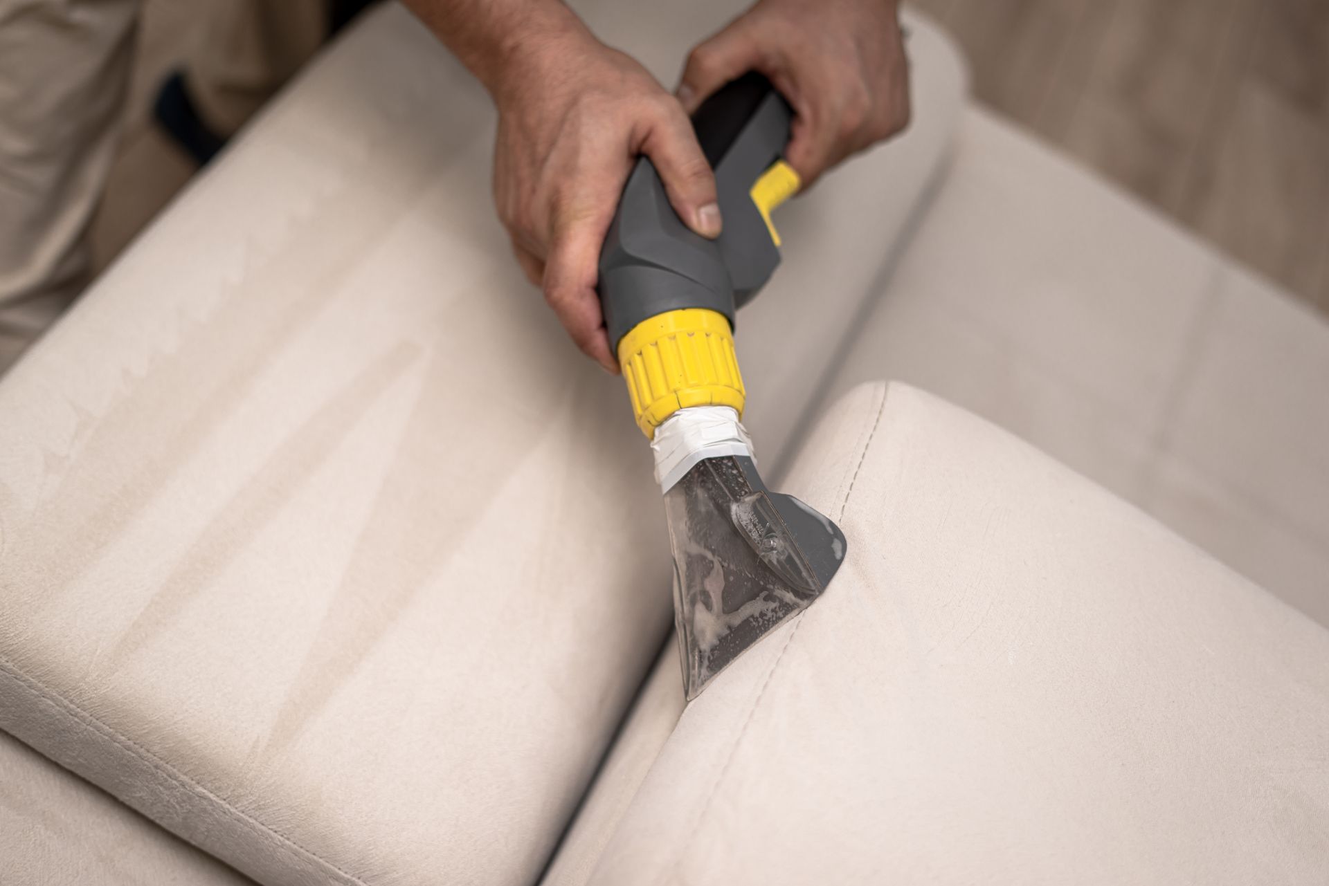 Person cleaning a light-colored sofa with a handheld upholstery cleaner; close-up view.
