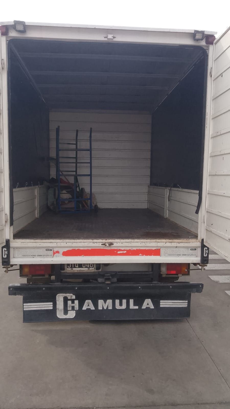 Tres operarios de mudanzas descargan muebles y cajas de una furgoneta blanca en una calle de las afueras.