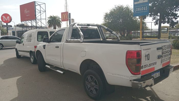 Camioneta blanca estacionada al costado de la carretera cerca de vallas publicitarias, con otra furgoneta blanca a su lado.