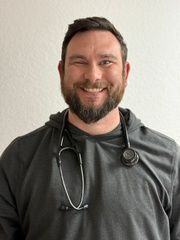 Man with a beard, stethoscope around his neck, winking and smiling, wearing a gray hooded shirt.