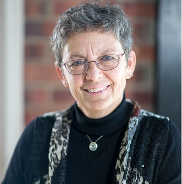 Woman with gray hair and glasses smiles. Black turtleneck, patterned vest. Brick wall background.