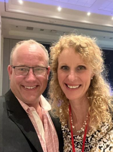 Smiling man and woman in a room; the man has glasses and a suit, woman with curly hair, both smiling at the camera.