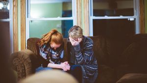 Two people, a redhead and a blonde, sit on a couch, heads bowed, appearing distressed in a dimly lit room.