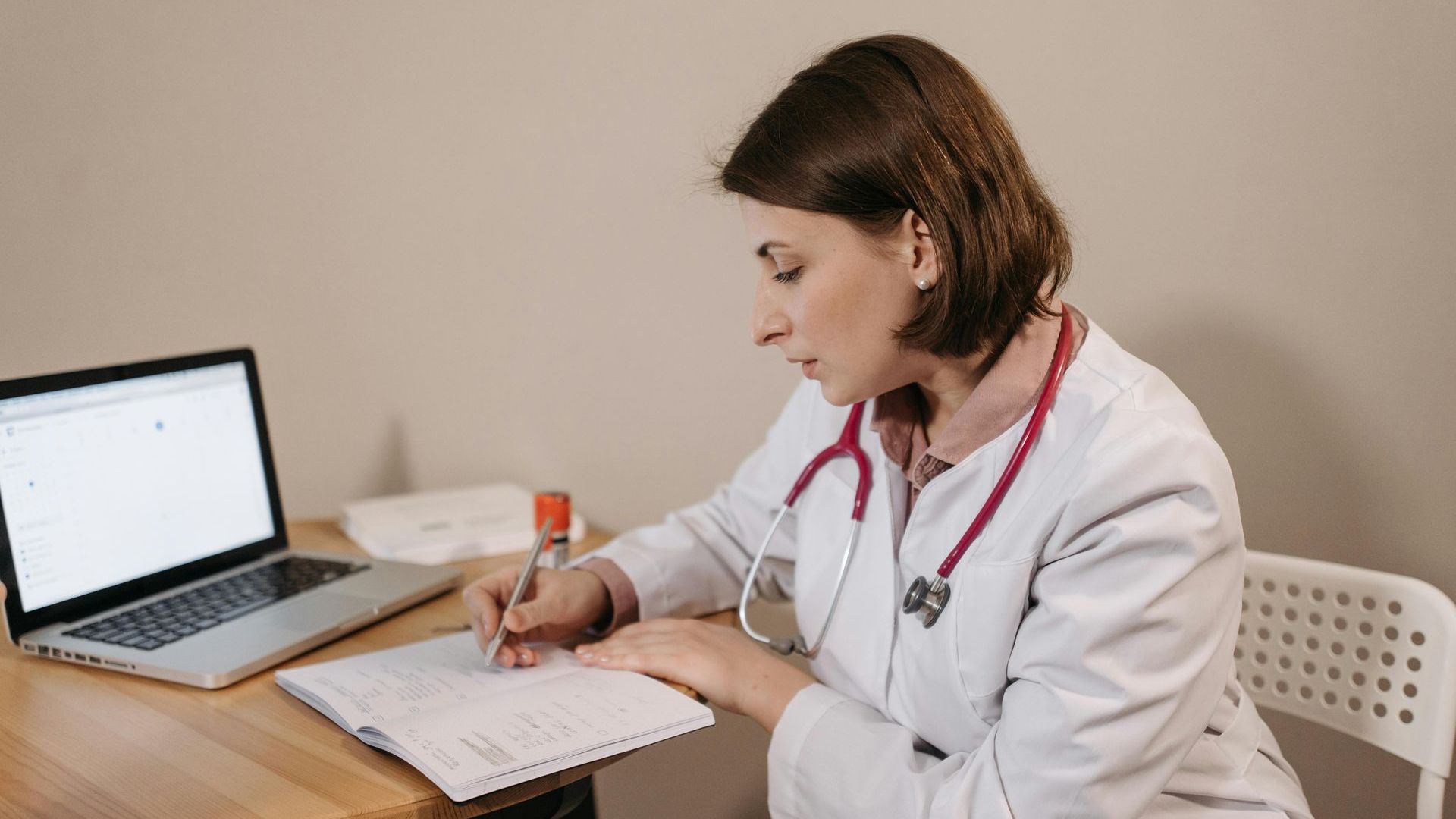 Female doctor in lab coat, stethoscope, writing at desk with laptop.