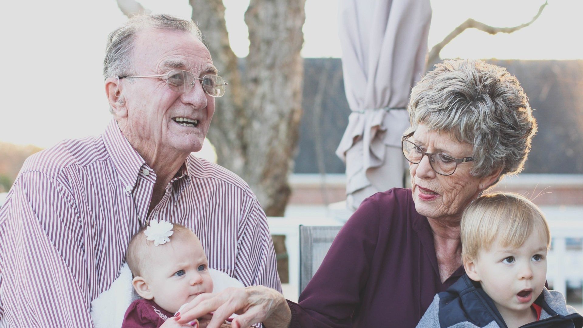 Grandparents with two young children outdoors; man in striped shirt, woman in purple, sunny.