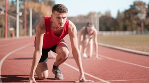 Man in red tank top in starting position on a track, focused expression, other runner blurred in background.