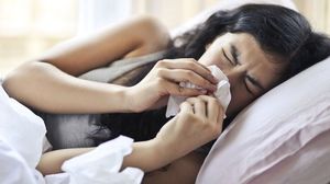 Woman in bed, blowing her nose with tissue, looking unwell.