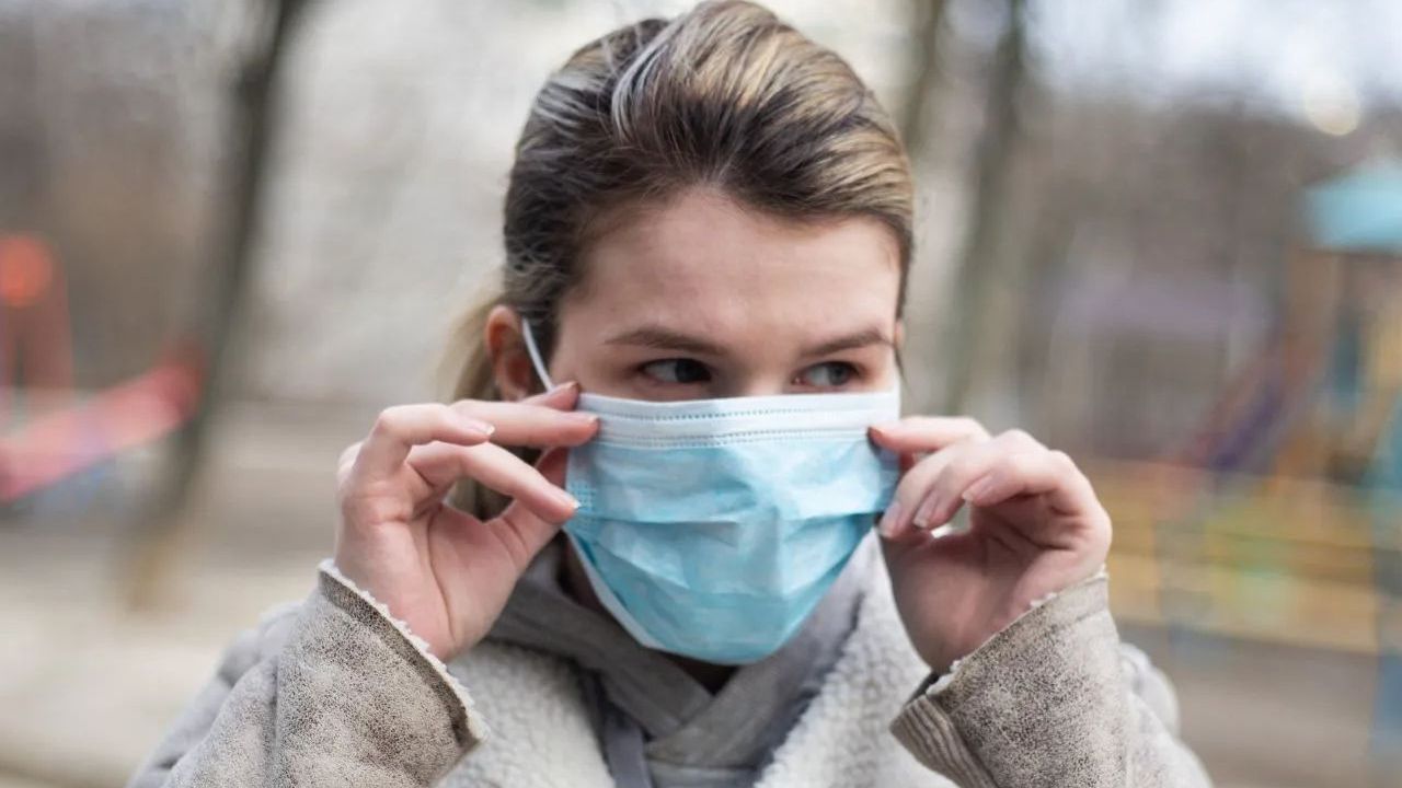Woman adjusting a blue surgical mask outdoors, with a neutral expression.