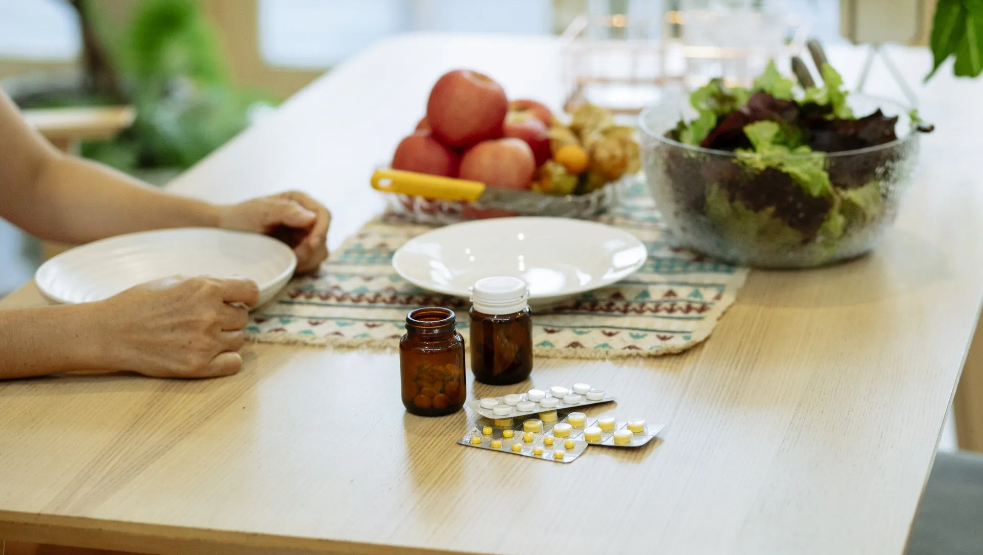 Person at table with pills, fruit, and salad. Hands reach for a white plate.