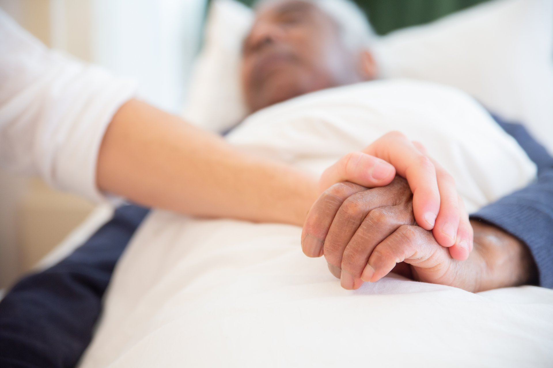 A person in bed holds hands with another person. Focus on hands, soft lighting.