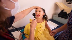Child with mouth open being examined by a doctor with an otoscope. Parent holds child's hands.