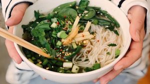 Person holding a bowl of noodles with greens, chopsticks.