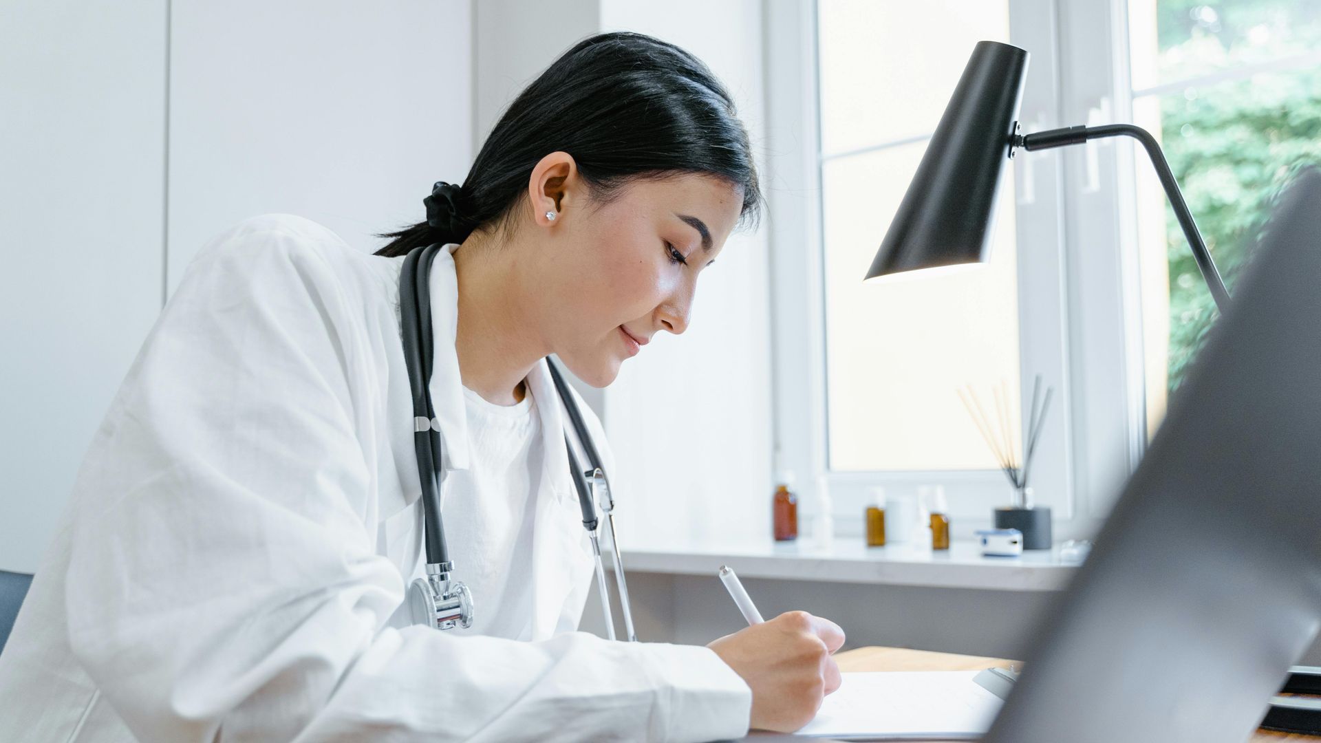 Female doctor in white coat writing at a desk, looking down.