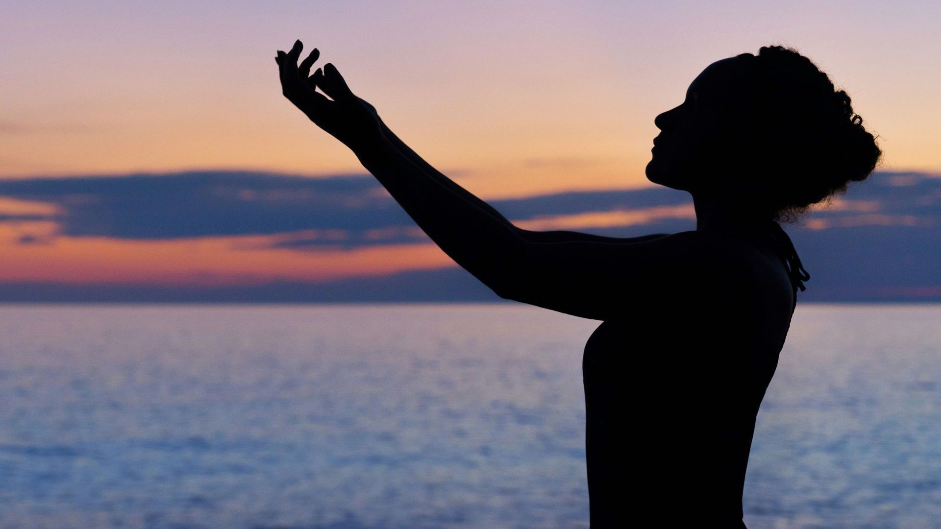 Silhouette of woman with arms raised toward a colorful sunset over water.