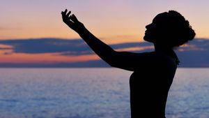 Silhouette of a woman with arms raised to the sky at sunset over the ocean.