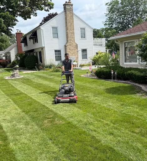 Person mowing a striped lawn in front of a white house on a sunny day