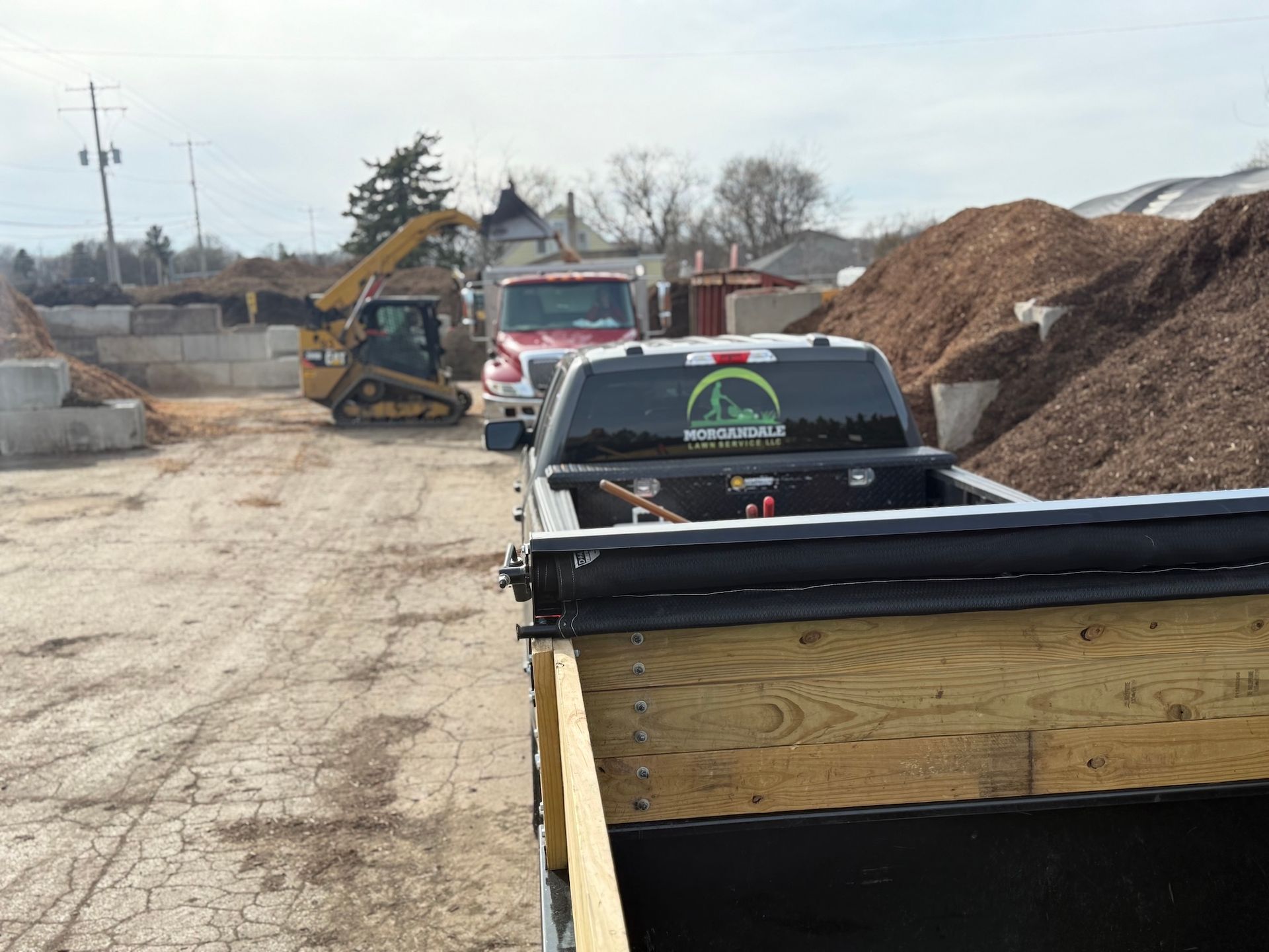 Construction site with skid steer, wood chips, and a pickup truck beside a wood-sided trailer